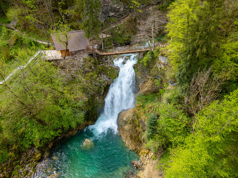 Aerial view of Slap Sum waterfall with a wooden bridge and a small hut surrounded by lush green forest in Zgornja Besnica, Kranj, Slovenia.