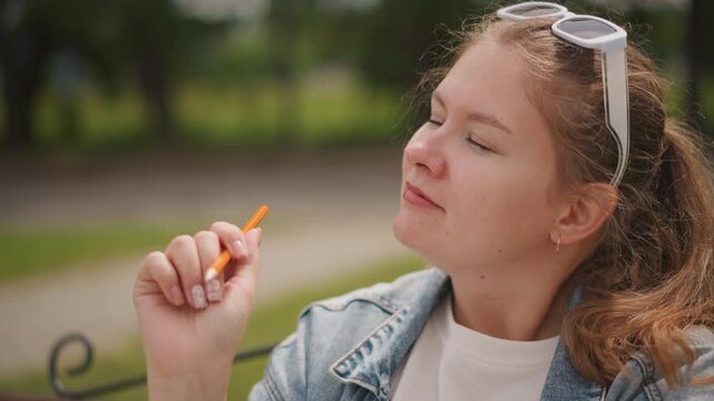 Young lady analyzing ecigarette outdoors. Teen girl observing vaping device on park bench. Candid portrait of youthful woman in denim jacket examining vape in park setting with soft background