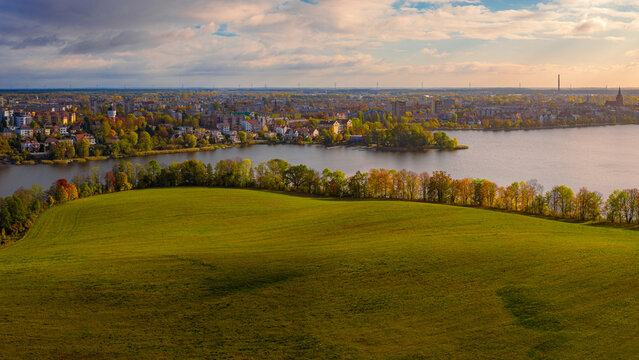 Aerial view of the city skyline across the lake with autumn trees and a green field in the foreground under a cloudy sky at sunset Elk, Warmian-Masurian Voivodeship, Poland.