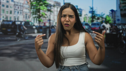 Fototapeta premium Hispanic woman points fingers at her chest with bare arms visible on a busy street; confusion doubt.