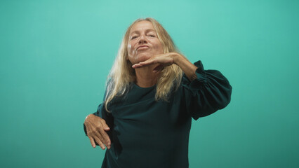 Senior blonde woman smiling and raising both hands with palms visible, dancing and swaying in a teal studio behind a seamless backdrop; joy celebration.