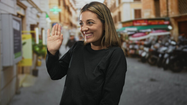 Young blonde woman waving open hand on a crowded city street market, smiling with eyes closed; happy greeting.