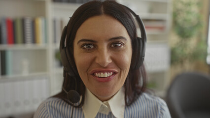 Woman wearing headset and microphone speaking and smiling, visible teeth, striped blouse, seated at desk inside office building with bookshelves and plant  warmth connection. © Krakenimages.com