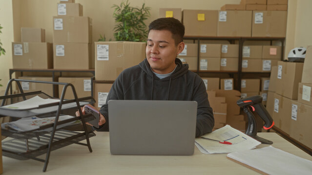 Man at laptop smiling and typing while glancing at phone amid stacked parcels in a building; small business contentment.