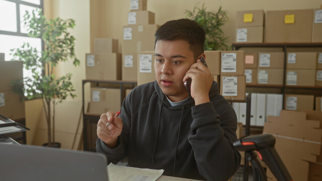 Man holding smartphone to ear and pen in hand, mouth slightly open, surrounded by stacked parcel boxes in building office; focused workday.
