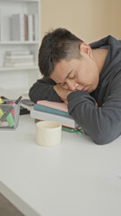 Obraz premium Man sleeping with head on stacked books at a library desk in a building, hoodie on, coffee mug and papers beside him; fatigue.