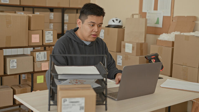 Man typing on laptop at a packing table surrounded by cardboard boxes and barcode scanner in a small business building; small business focus.