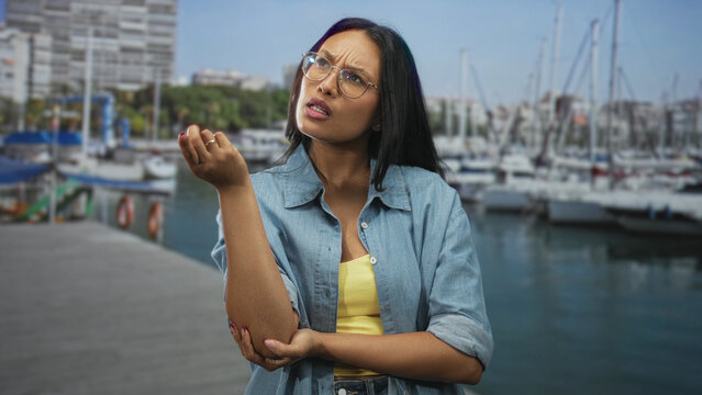 Woman wearing glasses points finger and holds forearm while standing on a marina pier near moored boats; disapproval assertive communication.