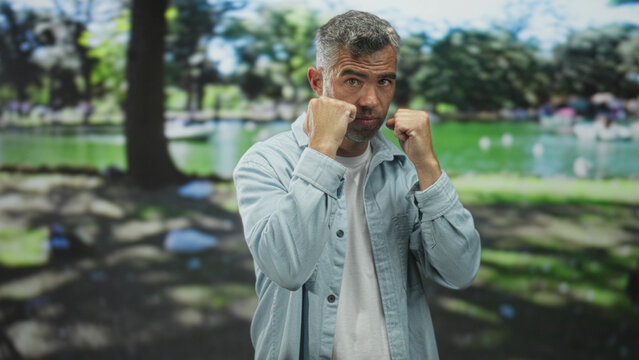 Man with grey hair shadowboxing with raised fists and guarded stance in a forest setting near a pond, wearing light denim shirt; training focus.