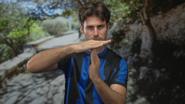 Man, young hispanic soccer fan, with hands forming timeout sign in forest path, wearing blue and black soccer jersey and staring at camera; team determination.
