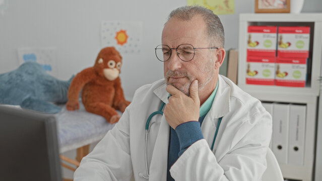 Man doctor, middle age, wearing glasses and stethoscope, hand on chin at computer in pediatric clinic with stuffed monkey on exam couch; thoughtful concern.