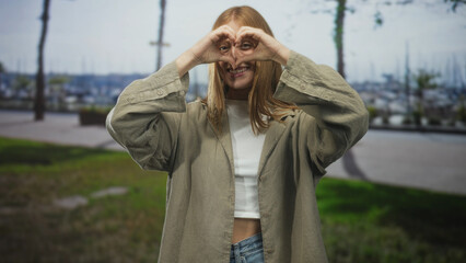 Fototapeta premium Young redhead woman forms heart with hands over eyes on a street by a marina pier, smiling in casual jacket and jeans; joyful playfulness.