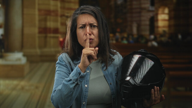 Middle age woman holding a glossy black motorcycle helmet, finger to lips for silence while standing inside a church nave with wooden pews and stone columns; quiet contemplation.