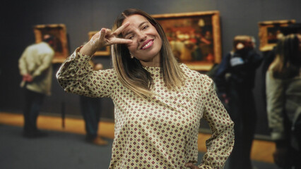Fototapeta premium Middleaged woman flashing peace sign with hand in museum building among framed paintings and visitors; playful confidence.