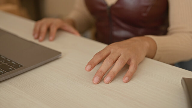 Woman hand hovering over light wood desk beside laptop in studio, fingers slightly splayed and poised above surface; focused productivity.