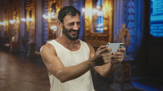Bearded middleaged man holds smartphone with both hands, smiling and showing bare forearms in a historic building gallery; joyful connection.