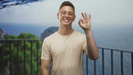 Obraz premium Man smiling makes ok sign with right hand on balcony railing at building overlooking sea cliff and trees; confidence relaxation.