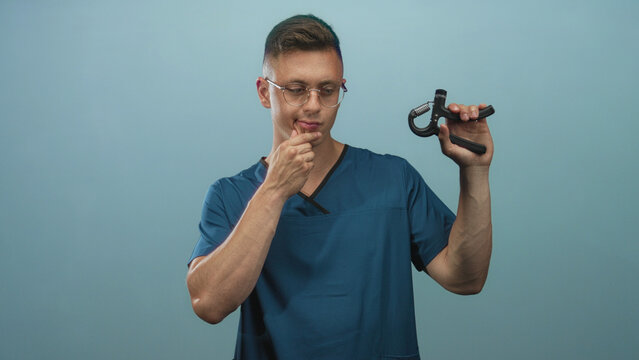 Young blond man physiotherapist in blue medical scrubs holds a handgrip exerciser in one hand while touching his chin in studio; thoughtful assessment.