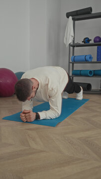 Man clasping hands and holding elbow plank on blue yoga mat beside exercise ball and shelf in studio; determination core wellness.