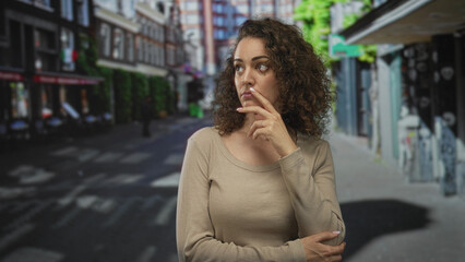 Obraz premium Woman with hand on chin at a city street crosswalk, neutral expression and folded arm across body, visible ring on finger; quiet contemplation.