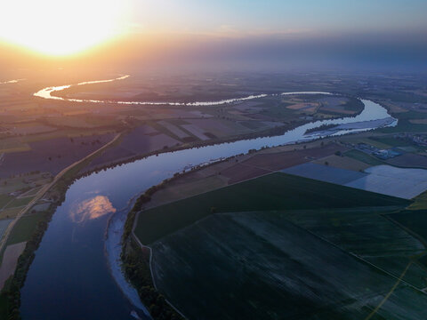 Aerial view of the winding Fiume Po flowing through agricultural fields under a golden sunset sky in Cotrebbia Vecchia, Emilia-Romagna, Italy.