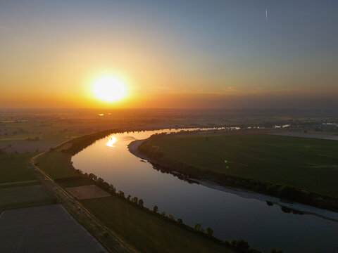Aerial view of the winding Fiume Po at sunset with golden light reflecting on the water surface and surrounding green fields in Cotrebbia Vecchia, Emilia-Romagna, Italy.