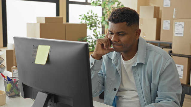 Man using computer with hand on chin and rubbing temples, surrounded by stacked parcel boxes and shipping labels inside a building; frustration.
