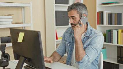 Naklejka premium Man holding smartphone to ear and pointing at computer monitor while seated at desk in office building, wearing denim jacket and beard visible; focused productivity.