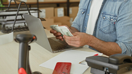 Obraz premium Man counting cash with bare hands beside laptop and packages at a parcel desk in building; small business hustle determination.