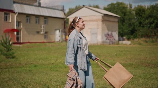 Woman strolling past suburban houses. Shopper on light errands with tote and patterned bag, denim jacket and jeans, headband, sunny afternoon, grassy yard and quiet neighborhood, easy pace