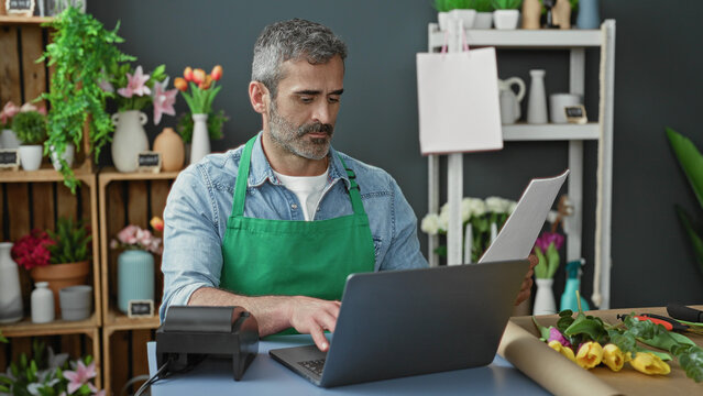 Man florist in green apron typing on laptop and holding order sheet with hands among flowers and packing tools at a shop counter inside a building; small business focus.