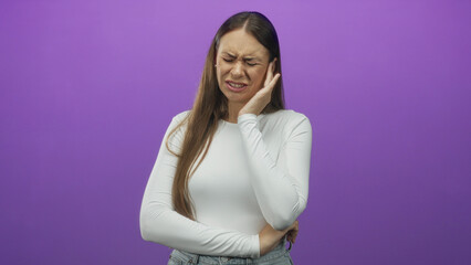 Obraz premium Woman holding her ear with her hand, eyes closed and grimacing while wearing a white long sleeve top in a purple studio; discomfort.