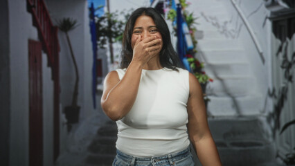Obraz premium Young hispanic woman in white sleeveless top and jeans covers mouth with hand while smiling on a narrow street by white steps; surprise delight.