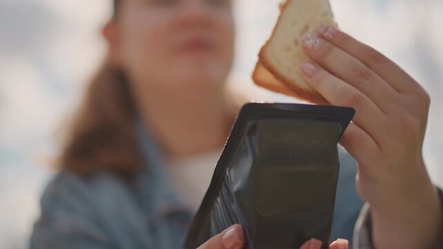 Young white woman opening sandwich box briskly, denim jacket, sunlit field, closeup hands pulling black container lid, revealing toasted sandwich with jam, candid snack moment, relaxed picnic vibe