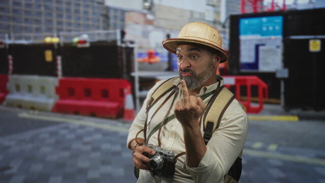 Man middle aged with grey beard and pith helmet shows middle finger while holding a vintage camera and backpack on a street construction site; defiance.