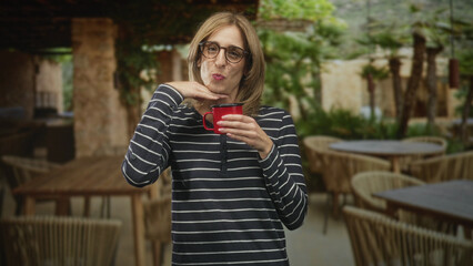 Fototapeta premium Woman holding red mug with hand under chin gesture, wearing glasses and striped shirt in a building cafe patio; playful delight.