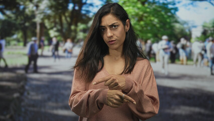 Fototapeta premium Woman in pink sweater points finger downward toward pavement on a crowded park walkway; doubt concern.