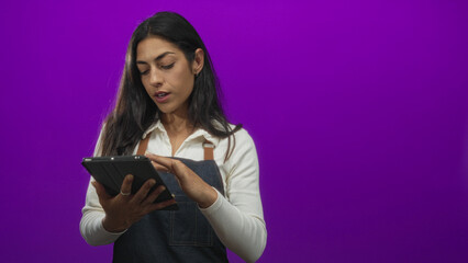 Naklejka premium Woman in a denim apron and white shirt tapping a tablet with fingers while reviewing an order list in a purple studio; concentration productivity.