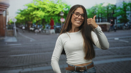 Fototapeta premium Young hispanic woman wearing glasses smiles and gives thumbs up with bare hands on an urban street with trees and bikes; confidence positivity.