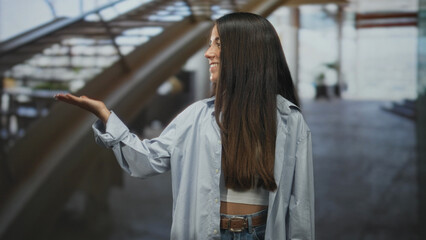 Obraz premium Young hispanic woman smiling with open palm gesture in airport terminal, wearing white crop top and blue shirt; friendly confidence.
