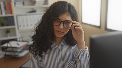 Woman adjusting glasses with hand, slight smile while seated at desk by monitor and bookshelves in an office building  confidence. © Krakenimages.com