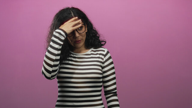 Woman touching forehead with hand while wearing glasses and striped top in pink studio; stress recovery.