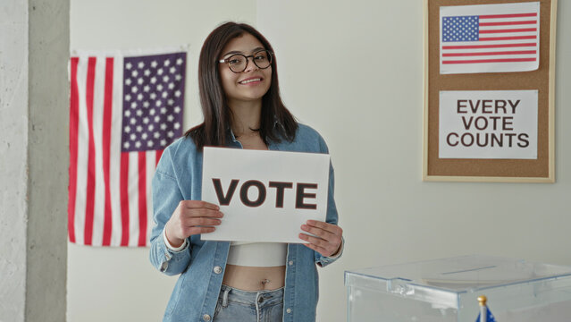 Young hispanic woman in denim jacket holding a vote sign with both hands beside a clear ballot box and us flag in a polling building; civic pride.