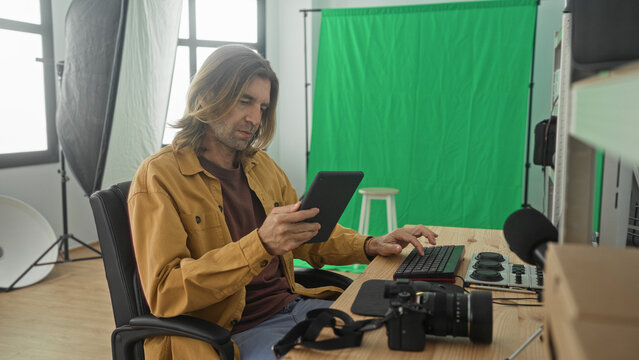 Man photographer holds tablet and types keyboard in studio with green screen and camera gear; focused creativity.