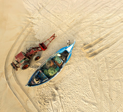 Aerial view of a traditional blue fishing boat and a red tractor on the sandy beach with tire tracks and a fisherman Praia de Mira, Coimbra, Portugal.