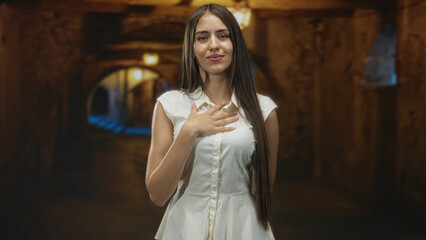 Fototapeta premium Young woman in white dress places hand on chest with soft smile and long hair visible in an arched underground stone corridor; serenity reflection.