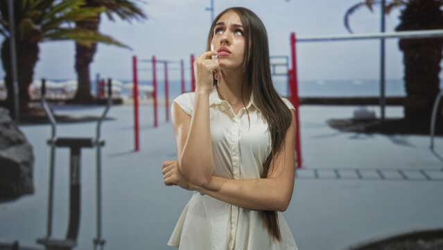 Young woman touching chin with finger and crossed arms at a beach outdoor gym by seaside pullup bars and palm trees visible; uncertainty.