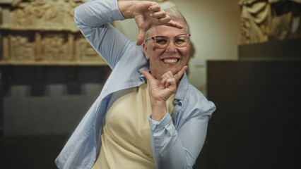 Obraz premium Middle age woman with glasses frames her face with hands and smiles while standing by museum exhibit in building; playful joy.