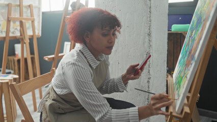 Fototapeta premium Woman painting colorful canvas with brush while holding smartphone and checking reference images in art studio; creative focus calm.