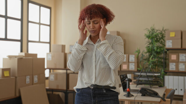 Woman pressing temples beside stacked cardboard boxes and a shipping parcel on a desk in a building, showing headache and fatigue; small business stress.